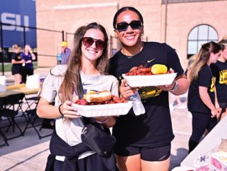 Two smiling women in sunglasses hold plates of food at an outdoor campus event, with a brick building and an LSU banner in the background.