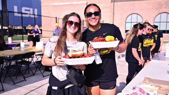 Two smiling women in sunglasses hold plates of food at an outdoor campus event, with a brick building and an LSU banner in the background.