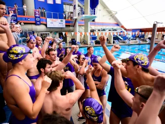 Swimmers in purple caps and suits huddled at pool edge, cheering and raising fists in celebration.