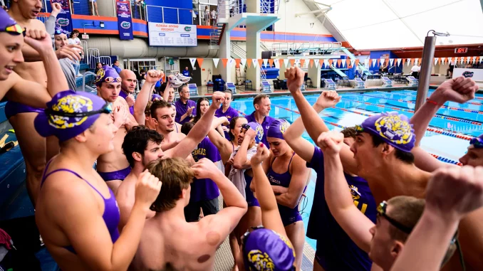 Swimmers in purple caps and suits huddled at pool edge, cheering and raising fists in celebration.