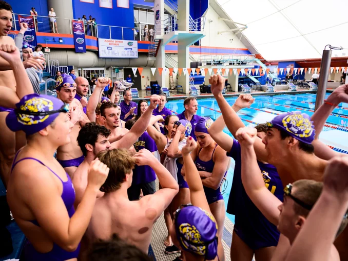 Swimmers in purple caps and suits huddled at pool edge, cheering and raising fists in celebration.