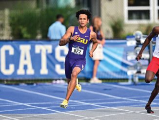 Two male sprinters race on a blue track; runner in purple LSU kit leads, bib 872, wearing yellow shoes.