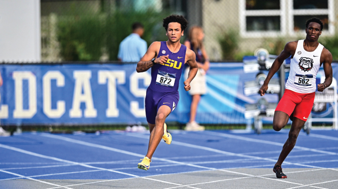 Two male sprinters race on a blue track; runner in purple LSU kit leads, bib 872, wearing yellow shoes.