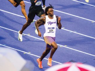 Sprinter in a white Tigers jersey running on a blue track with a bib number, chased by competitors in the background.