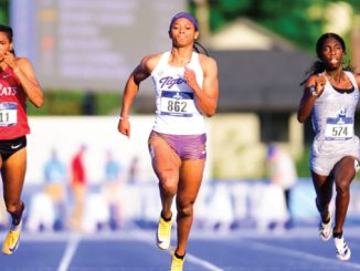 Three female sprinters race on a blue track in a stadium; bibs 211, 862, and 574 visible as they sprint mid-stride.