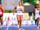 Three female sprinters race on a blue track in a stadium; bibs 211, 862, and 574 visible as they sprint mid-stride.