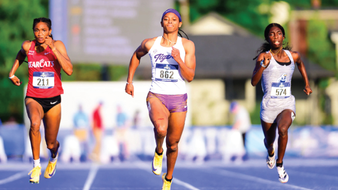 Three female sprinters race on a blue track in a stadium; bibs 211, 862, and 574 visible as they sprint mid-stride.