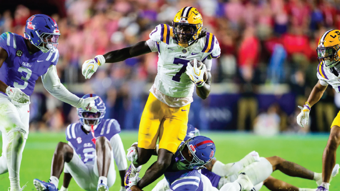 LSU player in white jersey with yellow pants runs with the ball while defenders in purple chase from behind on a college football field.