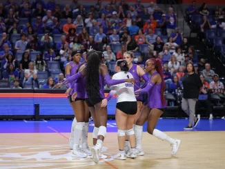 Group of female volleyball players in purple uniforms hug in a huddle on the court during a match you can see the crowd in the stands behind them.
