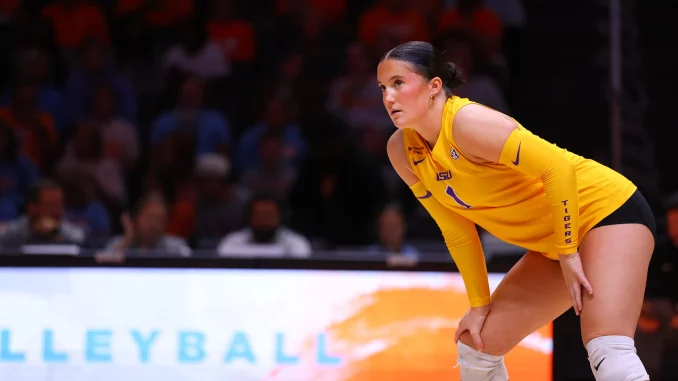Female volleyball player in a yellow LSU jersey crouches on the court, ready to receive the ball as spectators watch in the background.
