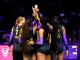 Women’s basketball team huddling with hands joined in a circle before a game, backs facing camera in black and purple uniforms.