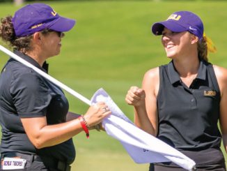 Two LSU female golfers in purple caps and black polos smile at each other while handling a white flag on a sunny golf course.