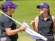 Two LSU female golfers in purple caps and black polos smile at each other while handling a white flag on a sunny golf course.