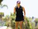 Female golfer in a black sleeveless polo and purple cap, walking with a golf club on a sunny course.