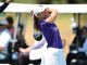 Female golfer in a purple shirt and white skirt mid-swing on a golf course, with spectators and golf carts in the background.