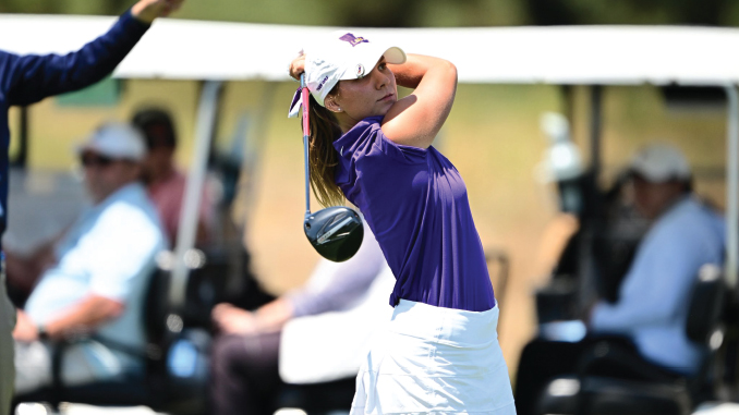 Female golfer in a purple shirt and white skirt mid-swing on a golf course, with spectators and golf carts in the background.