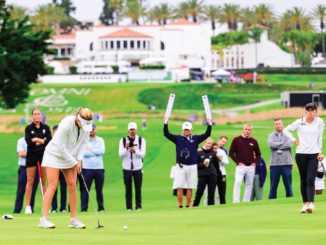 A female golfer lines up a putt on a sunny golf course while a crowd watches from the fairway behind her.