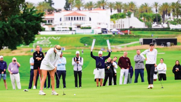 A female golfer lines up a putt on a sunny golf course while a crowd watches from the fairway behind her.