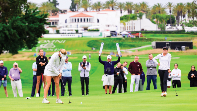 A female golfer lines up a putt on a sunny golf course while a crowd watches from the fairway behind her.