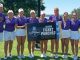 Eight LSU athletes in purple tops and white skirts pose on a sunny grassy field, holding a banner that reads '2026 Ticket Punched'.