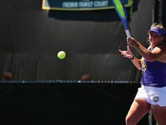 Female tennis player in purple top and white shorts about to hit a forehand as a ball flies toward her on an outdoor court