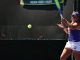 Female tennis player in purple top and white shorts about to hit a forehand as a ball flies toward her on an outdoor court