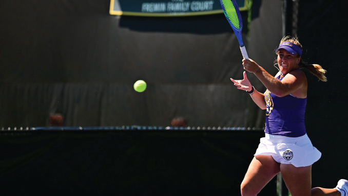 Female tennis player in purple top and white shorts about to hit a forehand as a ball flies toward her on an outdoor court