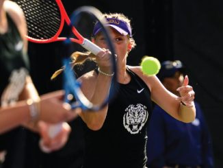 Female tennis player in a black tank top and purple visor swings a backhand as a bright tennis ball approaches, with another player blurred in the foreground.