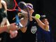 Female tennis player in a black tank top and purple visor swings a backhand as a bright tennis ball approaches, with another player blurred in the foreground.