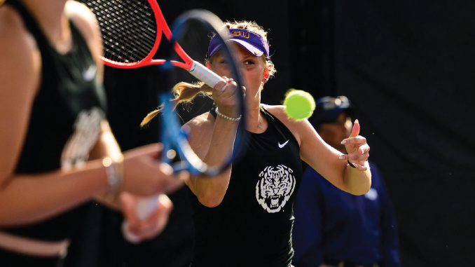 Female tennis player in a black tank top and purple visor swings a backhand as a bright tennis ball approaches, with another player blurred in the foreground.
