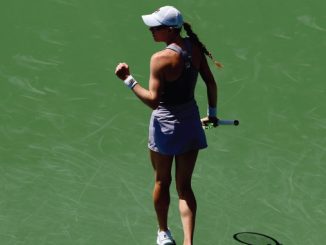 Female tennis player on a green court, seen from behind, wearing a cap and white skirt, holding a racket in her right hand.