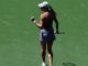 Female tennis player on a green court, seen from behind, wearing a cap and white skirt, holding a racket in her right hand.