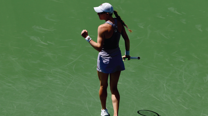 Female tennis player on a green court, seen from behind, wearing a cap and white skirt, holding a racket in her right hand.