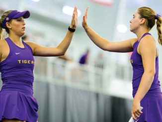 Two female athletes in purple uniforms high-five each other during practice in an indoor sports facility.