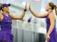 Two female athletes in purple uniforms high-five each other during practice in an indoor sports facility.