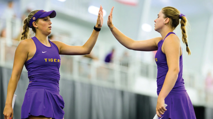 Two female athletes in purple uniforms high-five each other during practice in an indoor sports facility.