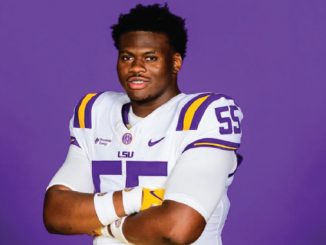 LSU football player in white jersey with purple and gold accents, arms crossed against a purple background.