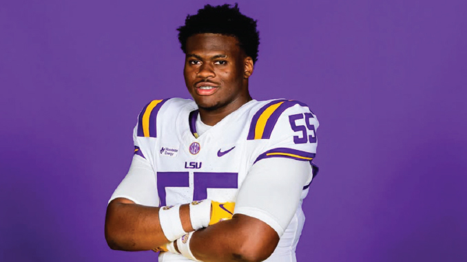 LSU football player in white jersey with purple and gold accents, arms crossed against a purple background.