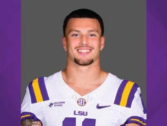 Smiling LSU football player in white jersey with purple-gold stripes, head-and-shoulders portrait against gray background