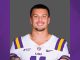 Smiling LSU football player in white jersey with purple-gold stripes, head-and-shoulders portrait against gray background