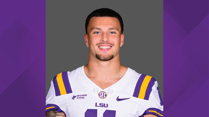 Smiling LSU football player in white jersey with purple-gold stripes, head-and-shoulders portrait against gray background