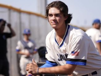 Young baseball player in a white jersey with blue trim leans on a railing during practice, USA flag patch on the sleeve, focused expression