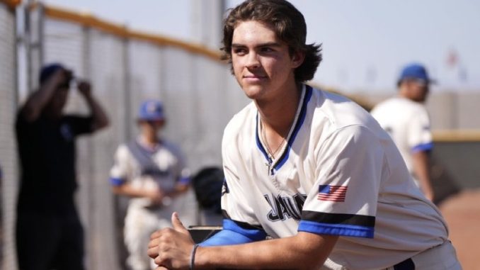 Young baseball player in a white jersey with blue trim leans on a railing during practice, USA flag patch on the sleeve, focused expression
