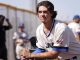 Young baseball player in a white jersey with blue trim leans on a railing during practice, USA flag patch on the sleeve, focused expression