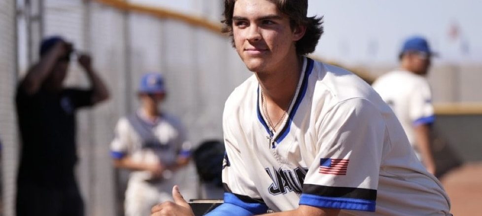 Young baseball player in a white jersey with blue trim leans on a railing during practice, USA flag patch on the sleeve, focused expression