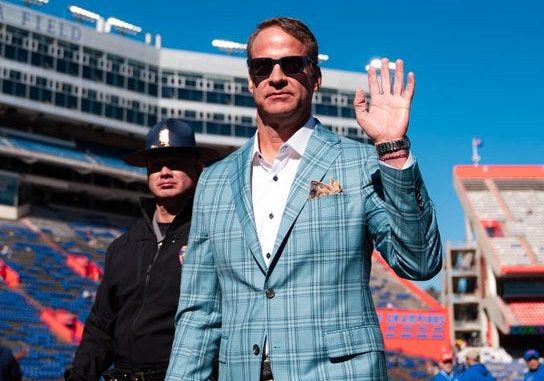 Man in a blue plaid blazer and sunglasses waves at the camera on a football field, with a security guard behind him in a stadium under a clear sky.