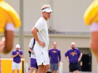 Coach in white polo and visor watches practice with hands on hips as players in purple and gold uniforms train in the background.