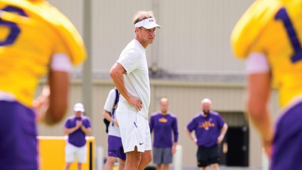 Coach in white polo and visor watches practice with hands on hips as players in purple and gold uniforms train in the background.