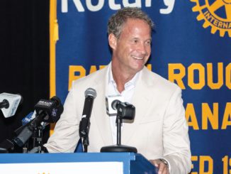 Man in a light beige suit smiles at a podium with multiple microphones, standing in front of a blue and yellow Rotary banner.