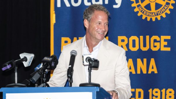 Man in a light beige suit smiles at a podium with multiple microphones, standing in front of a blue and yellow Rotary banner.